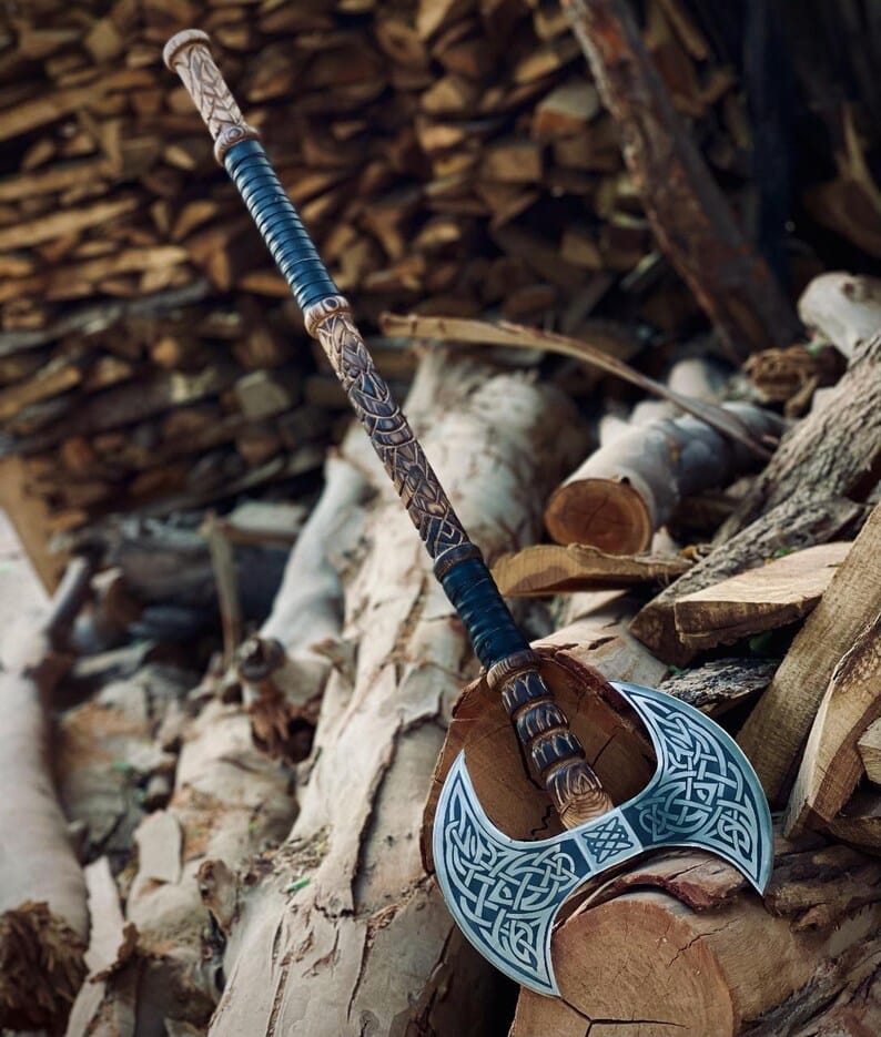 Viking Double Headed Axe with engraved Celtic knotwork and a hand-carved handle, resting on a pile of firewood.